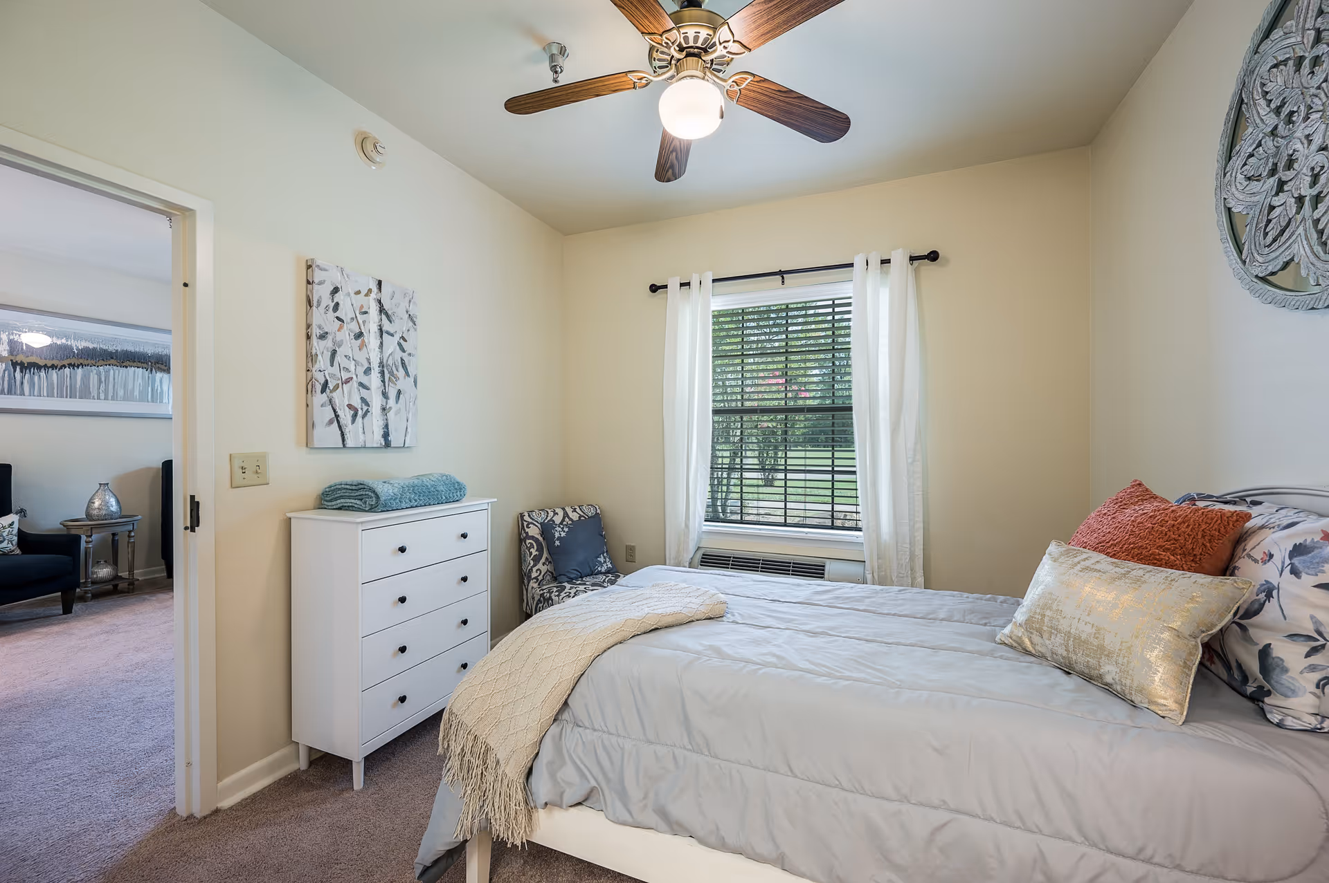 A cozy bedroom with a bed made up with gray bedding and decorative pillows, a white chest of drawers with a folded blanket on top, a patterned armchair, a window with white curtains, and a ceiling fan with a light. The room has beige walls and carpeted floor. An adjacent room with seating furniture is visible through an open door.
