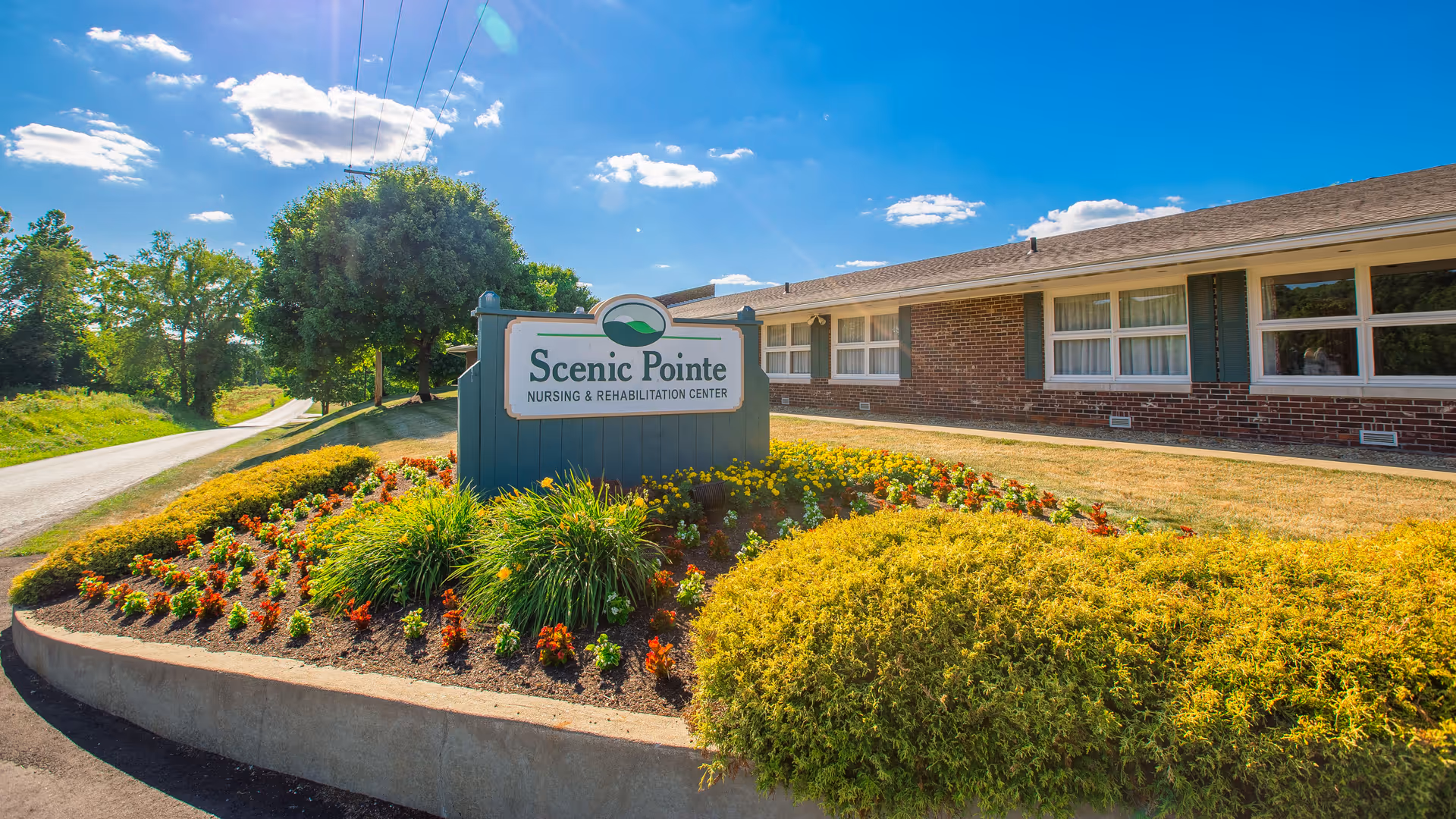 Exterior view of Scenic Pointe Nursing & Rehabilitation Center on a sunny day, showing a landscaped flower bed with colorful flowers and bushes in front of a brick building with multiple windows.