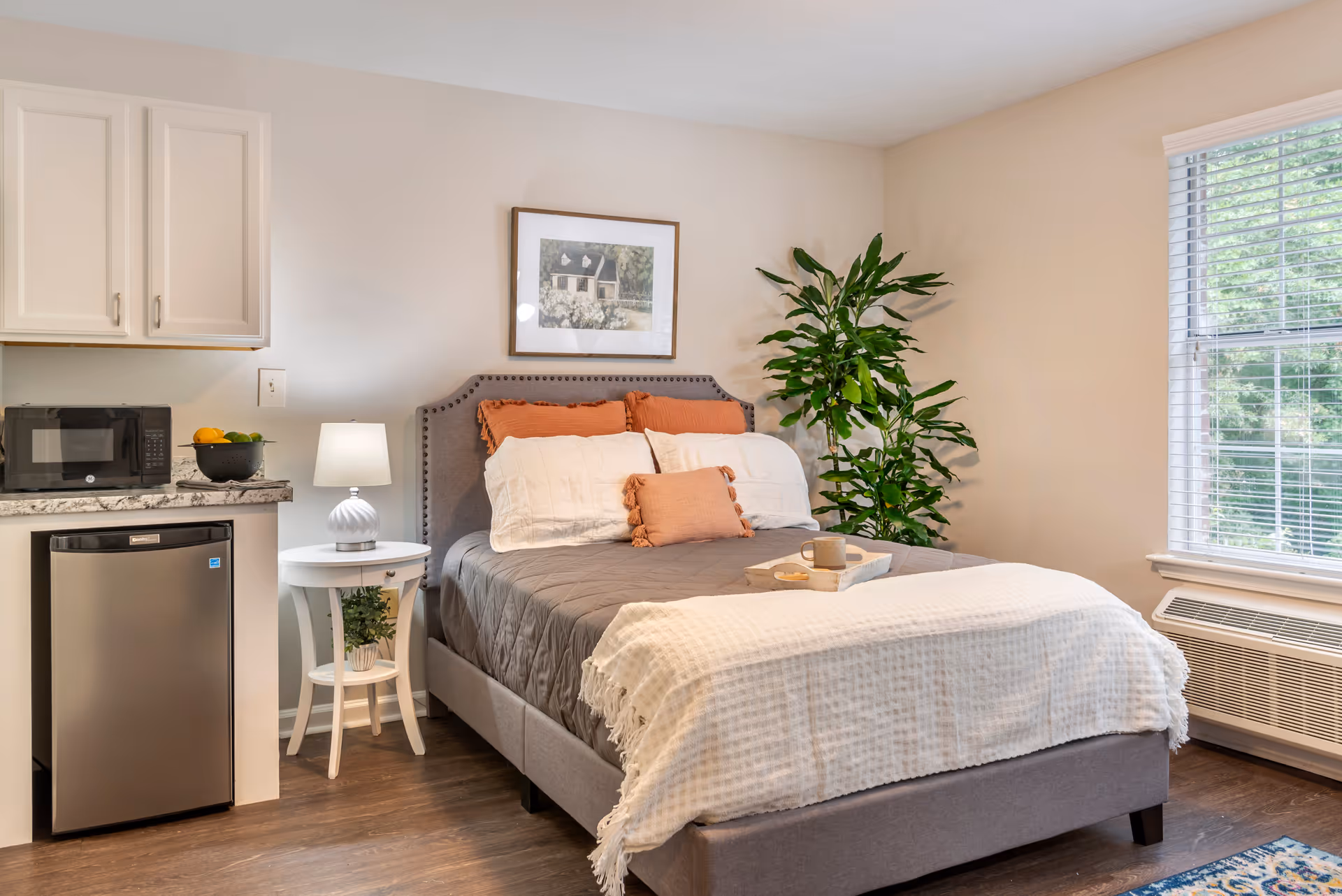 A cozy bedroom with a gray upholstered bed featuring white and orange pillows and a white throw blanket. Next to the bed is a small white round table with a lamp and a plant underneath. To the left, there is a small kitchenette area with a microwave, a bowl of fruit, and a mini refrigerator. A large window with blinds allows natural light to brighten the room, and a tall green plant is placed in the corner.
