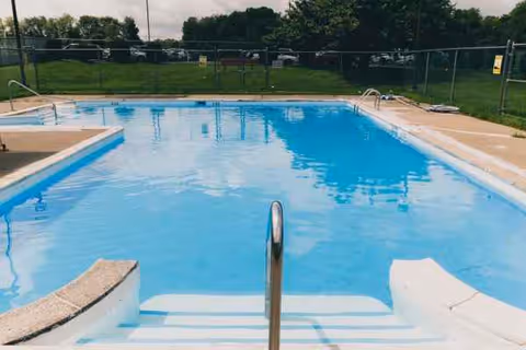 Outdoor swimming pool with a metal handrail, steps, and surrounding fence.