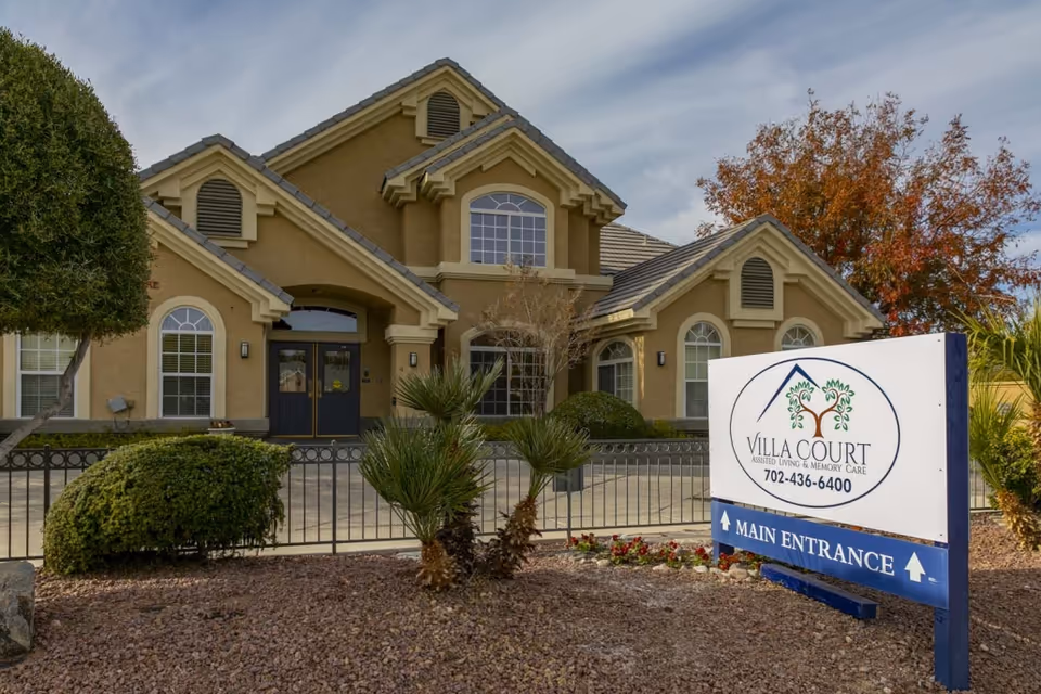 Front exterior of a two-story assisted living building with landscaping and a freestanding 'Villa Court' main entrance sign.