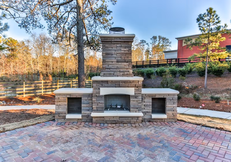 Stone outdoor fireplace and seating structure on a brick patio with trees and a building in the background.