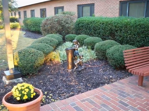 Brick-paved courtyard with a wooden bench, potted flowers, trimmed hedges, and a small decorative statue in a mulched garden bed in front of a brick building.