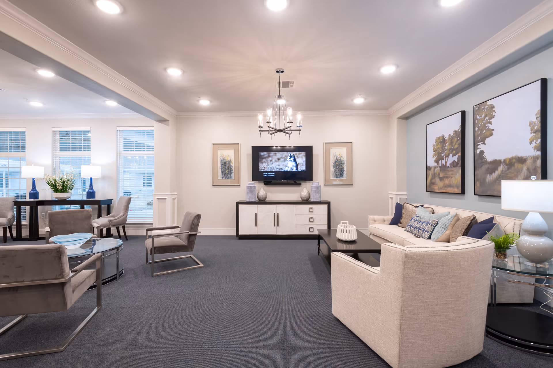 A well-lit living room area in Wesley House featuring a beige sofa with multiple cushions, two modern armchairs, a black coffee table, and a TV mounted on the wall above a white cabinet. The room has blue carpeting, light-colored walls with framed artwork, and a chandelier hanging from the ceiling. There is also a dining table with chairs near the windows on the left side.