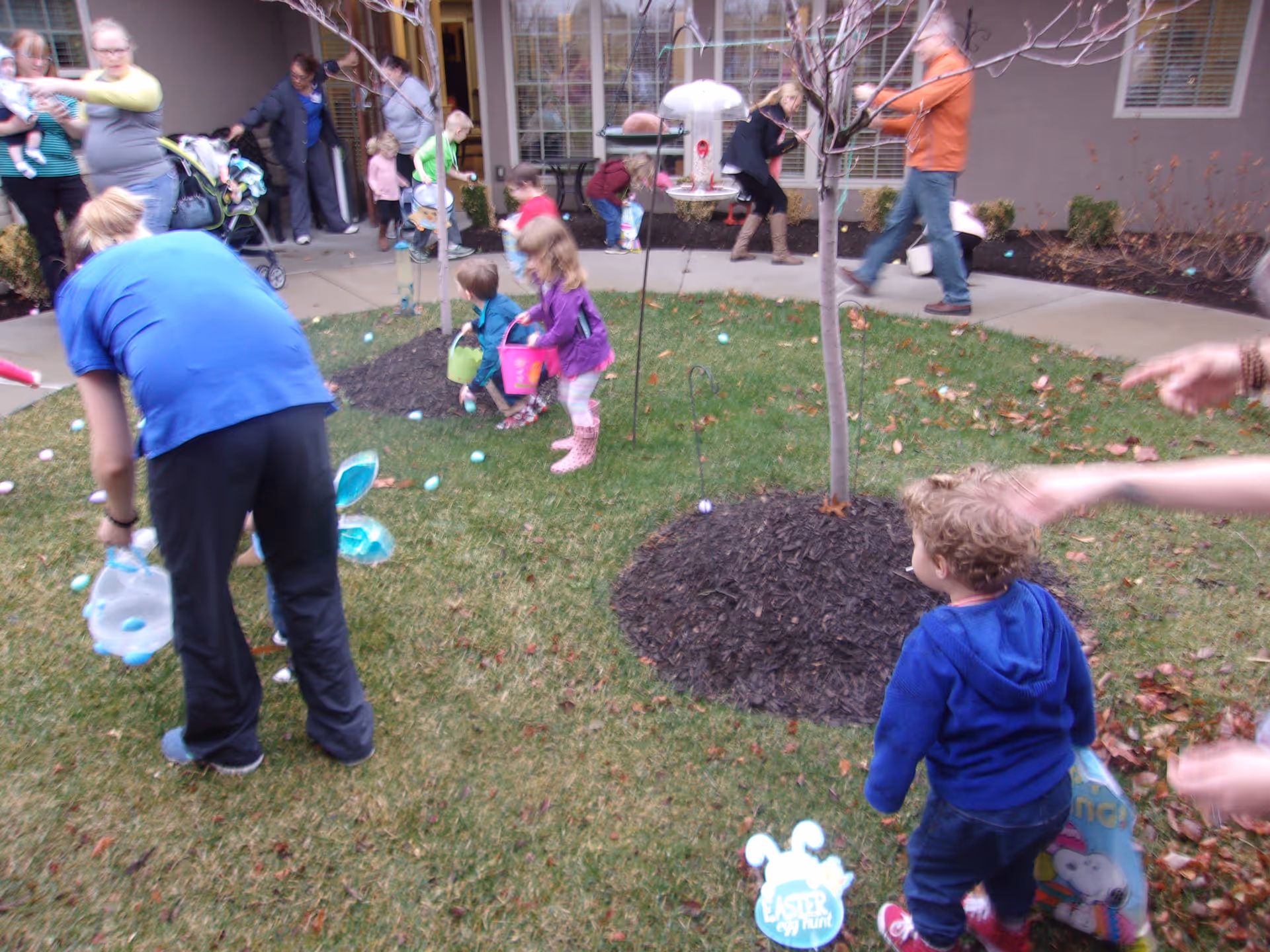 Children and adults participating in an Easter egg hunt on a grassy area with small trees and a building in the background. The children are holding baskets and collecting colorful eggs scattered on the ground.