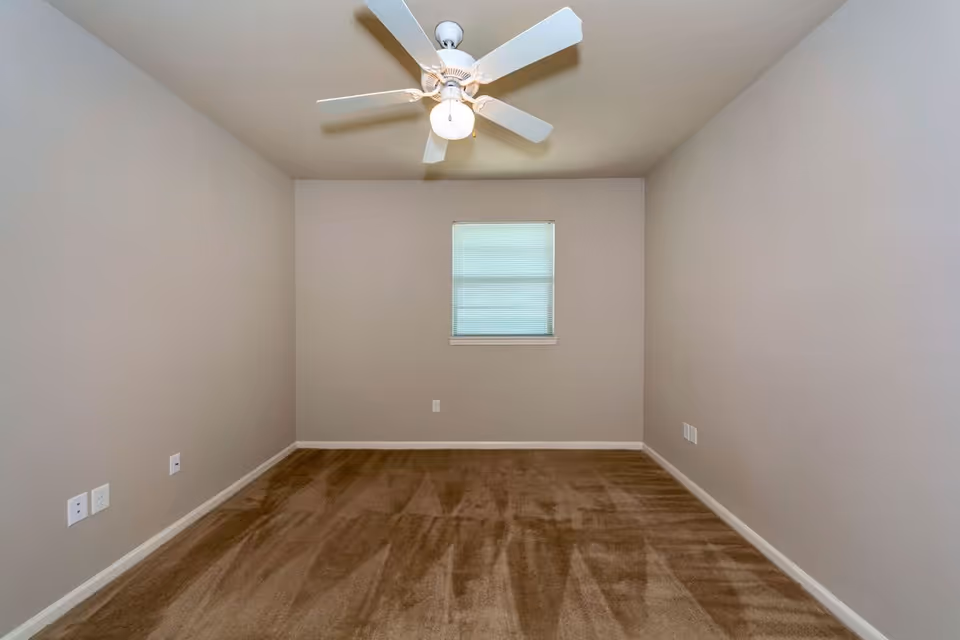 Empty bedroom with a ceiling fan, small window with a blind, beige walls, and brown carpet.