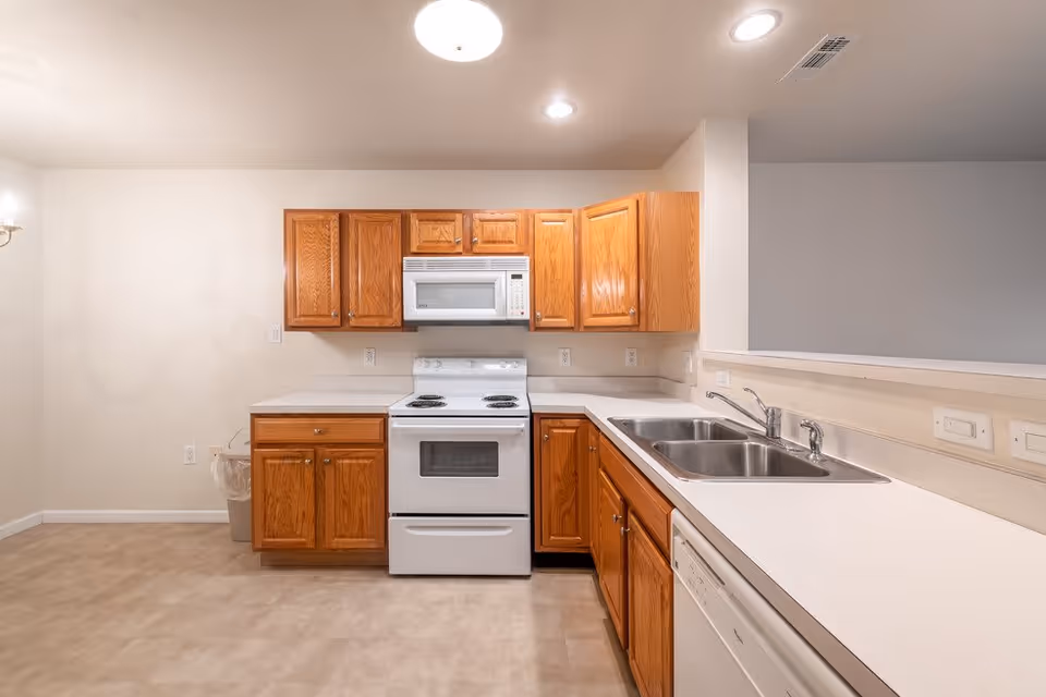 A clean kitchen with wooden cabinets, a white electric stove with oven, a white microwave above the stove, a double stainless steel sink, and a dishwasher. The countertops are white, and the floor has a light beige tile. The walls are painted white, and there are ceiling lights providing illumination.