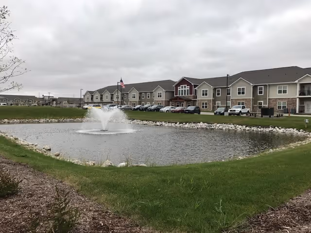 A pond with a water fountain in the center, surrounded by grass and landscaping. Behind the pond is a two-story residential building with multiple windows and parked cars in front. The sky is overcast.