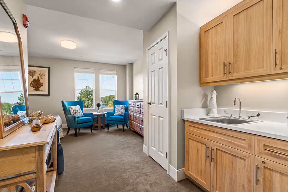Interior view of a senior living facility room featuring a small kitchenette with wooden cabinets and a sink on the right. In the background, there are two blue upholstered armchairs with patterned pillows, a small round wooden table between them, a wooden dresser, and two large windows letting in natural light. A wooden console table with decorative items and a large mirror is on the left side of the image.