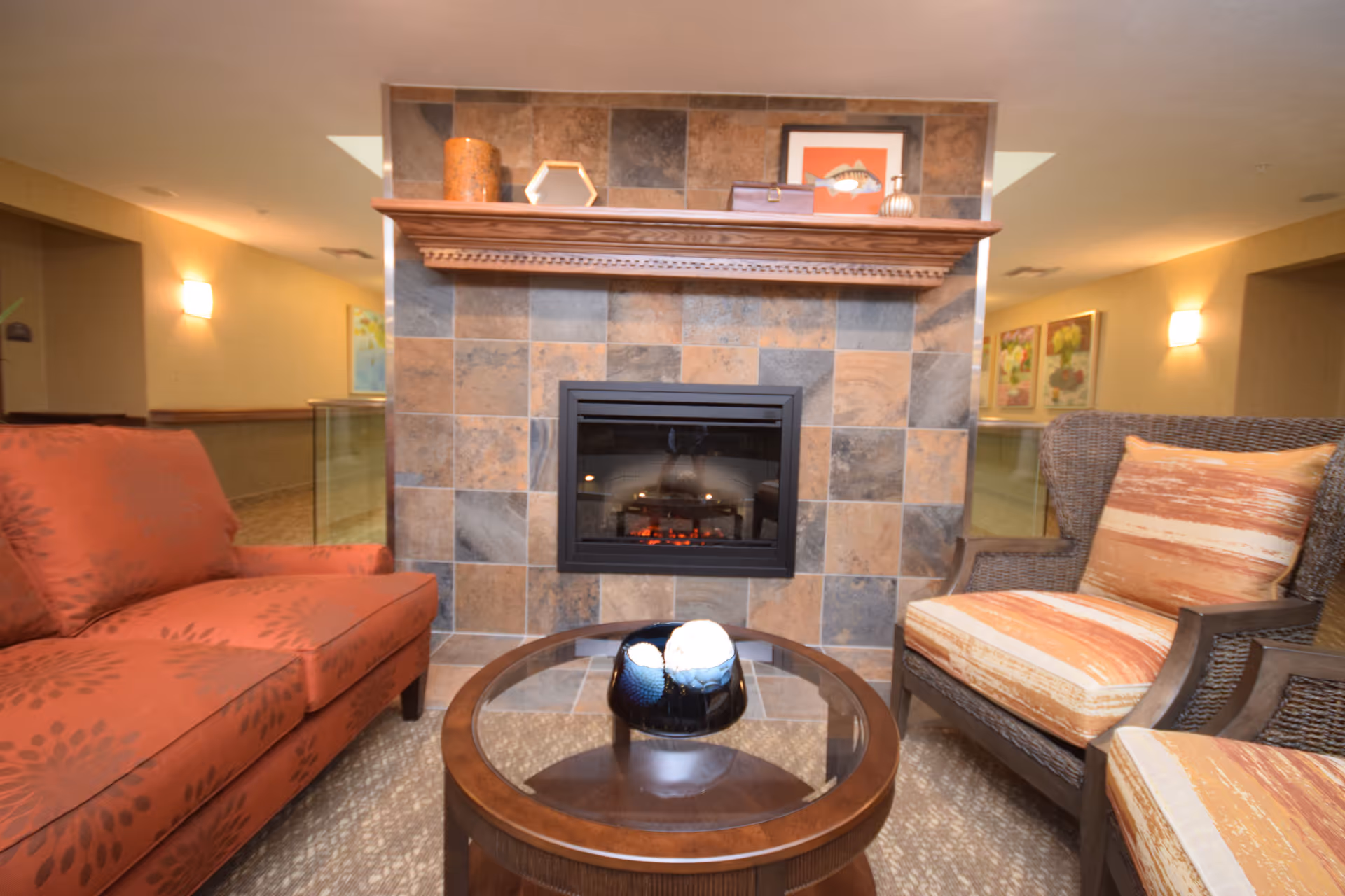 Cozy seating area in a retirement community featuring a tiled fireplace with a wooden mantel, a round glass-top coffee table with decorative items, an orange patterned sofa on the left, and a wicker armchair with orange and beige cushions on the right.