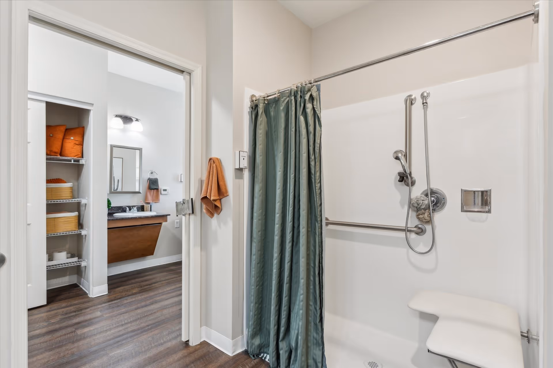 A bathroom with a walk-in shower featuring a green shower curtain, grab bars, a handheld showerhead, and a fold-down shower seat. Adjacent to the shower is a wall with a peach-colored towel hanging. Through an open doorway, a vanity with a sink, mirror, and light fixture is visible, along with shelving holding orange towels and baskets.