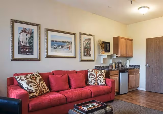 Interior view of a living area in a retirement community featuring a red sofa with patterned and solid cushions, three framed pictures on the wall above the sofa, a small kitchenette with wooden cabinets, a mini fridge, and a countertop with kitchen items, and a wooden door in the background.