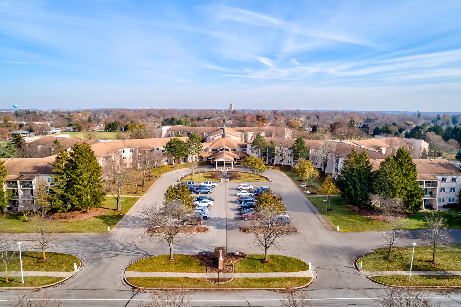 Aerial view of Holiday Winter Village senior living facility showing a large U-shaped building with a central entrance, surrounded by trees and parking lots with several cars parked. The sky is clear with some wispy clouds.