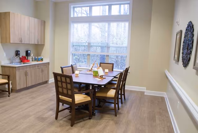 Small dining room with a wooden table and six chairs by a large window and a built-in kitchenette.