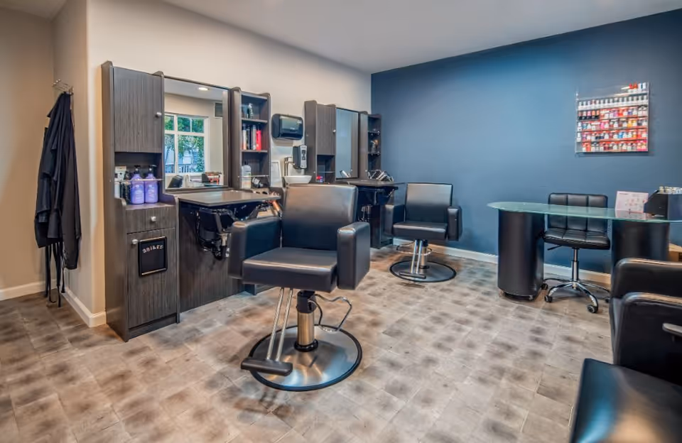 Interior view of a salon area in a senior living facility with two black salon chairs in front of mirrors and washbasins, a small desk with a chair, and a wall-mounted display of nail polish bottles on a blue accent wall.