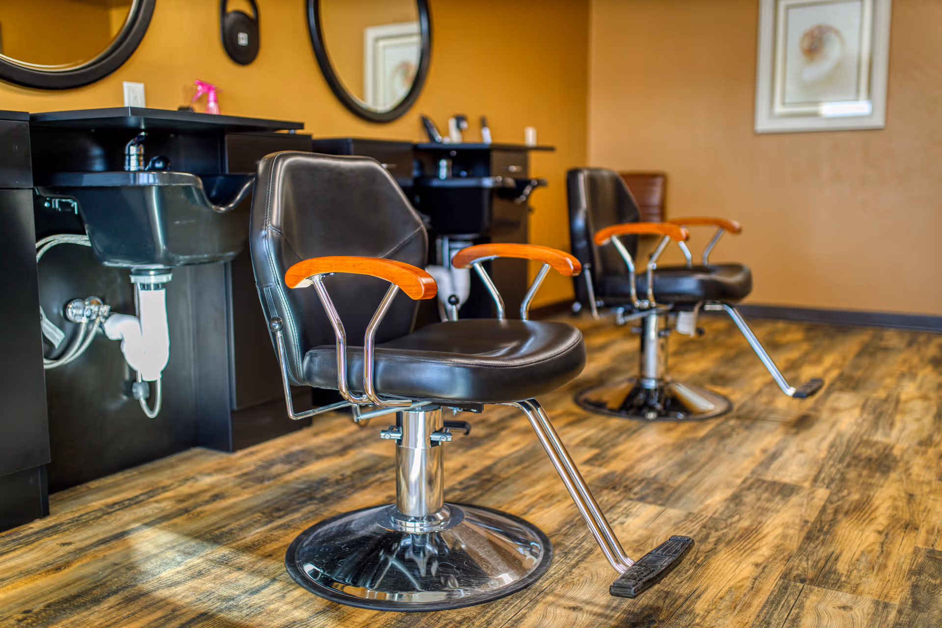 Interior view of a salon area with two black salon chairs featuring wooden armrests, positioned in front of black sinks and mirrors mounted on a mustard-colored wall, with wood-patterned flooring.