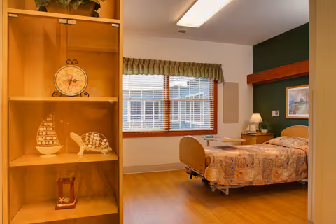 A cozy bedroom in a senior living facility featuring a single bed with a patterned bedspread, a wooden headboard, a bedside table with a lamp, a framed picture on the wall, a window with blinds and a valance, and a wooden shelving unit displaying decorative items including a clock, a model sailboat, and a turtle figurine.