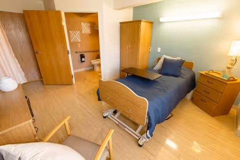 A well-lit bedroom in a skilled nursing facility featuring a single hospital-style bed with blue bedding and a small overbed table. The room includes wooden furniture such as a nightstand with a lamp, a dresser, a wardrobe, and a cushioned armchair. An open door reveals an adjacent bathroom with a toilet and wall decorations.