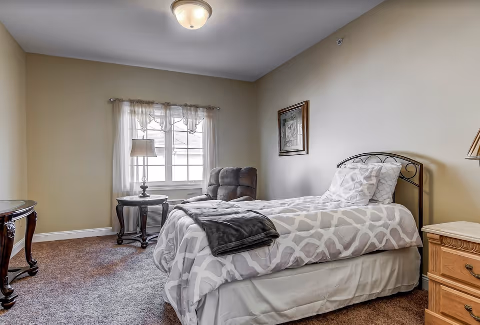 A cozy bedroom with a single bed covered in a white and gray patterned comforter and a dark gray blanket folded at the foot. Next to the bed is a wooden nightstand with a lamp. A comfortable armchair is positioned near a window with sheer white curtains, and a small round table with a lamp sits beside the chair. The walls are painted beige, and there is a framed picture hanging above the bed. The floor is carpeted in a brown tone.