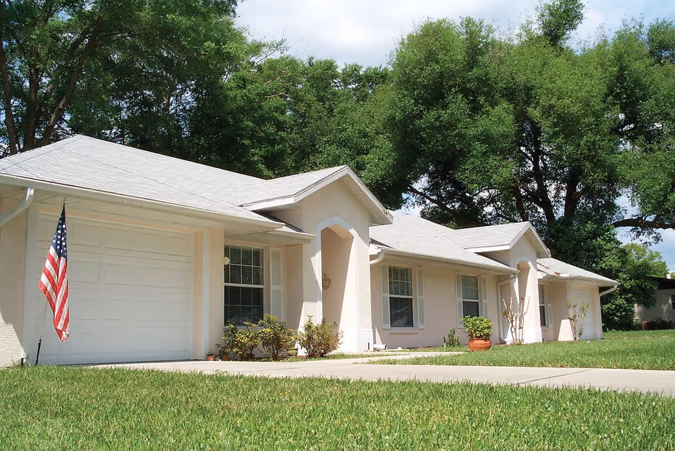 Front exterior of a single-story light-colored senior living building with a garage, American flag, windows, potted plants, and a grassy lawn.