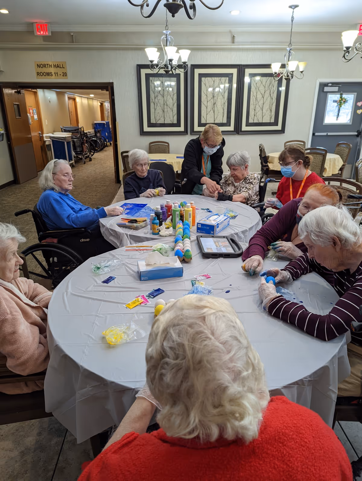 A group of elderly residents and staff seated around a table in a communal room painting and decorating eggs.
