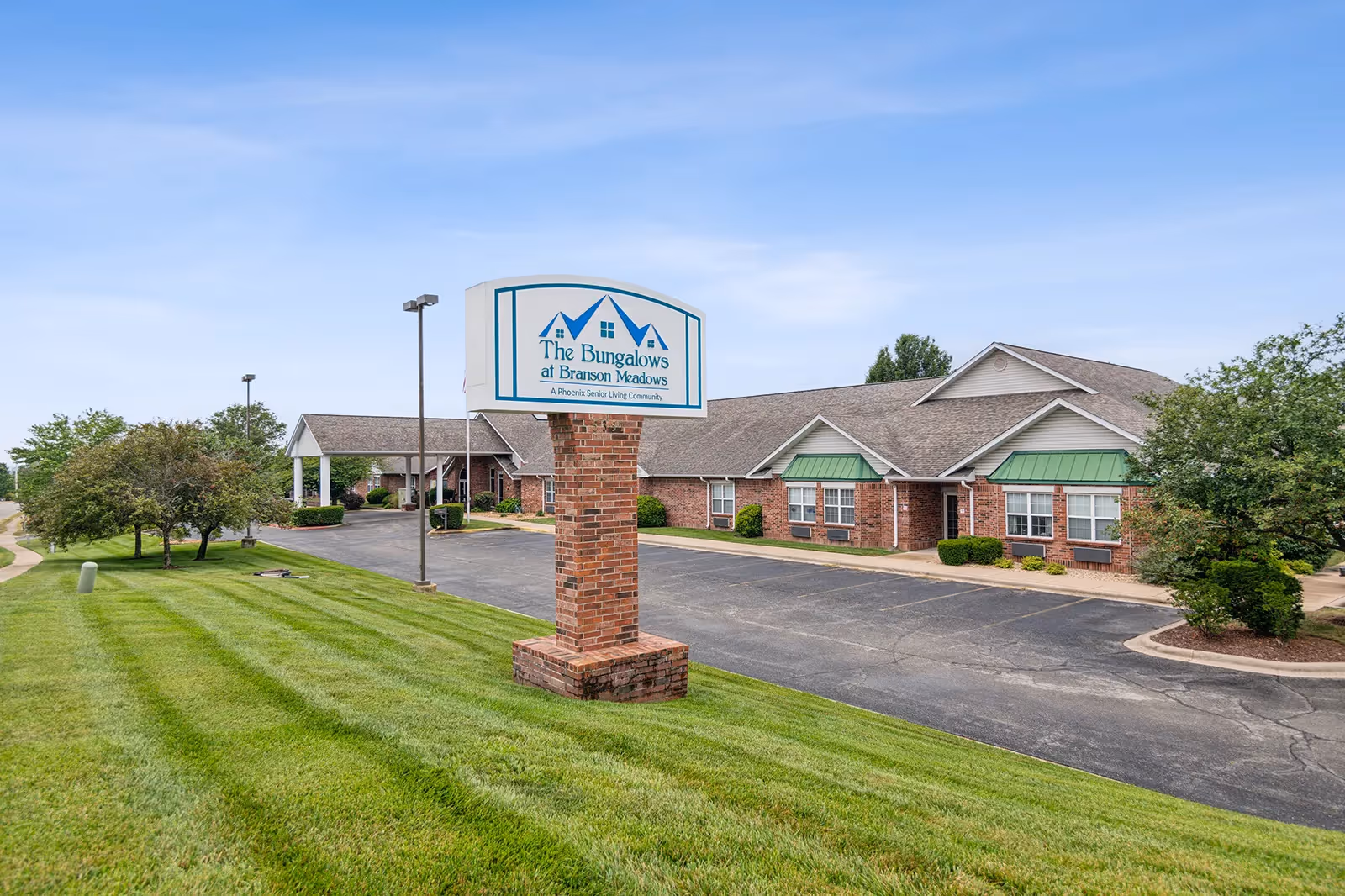 Exterior view of The Bungalows at Branson Meadows senior living community building with a brick signpost displaying the facility's name. The building has a brick facade with green awnings over the windows, surrounded by a well-maintained lawn and trees under a clear blue sky.