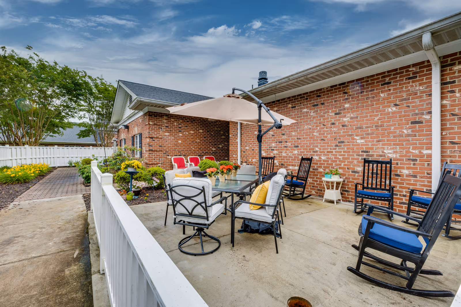 Outdoor patio area at Kerner Ridge Assisted Living featuring a glass-top table with cushioned chairs and a large umbrella, surrounded by several rocking chairs with blue cushions. The patio is adjacent to a red brick building and bordered by a white fence, with a garden and pathway visible on the left side under a partly cloudy sky.
