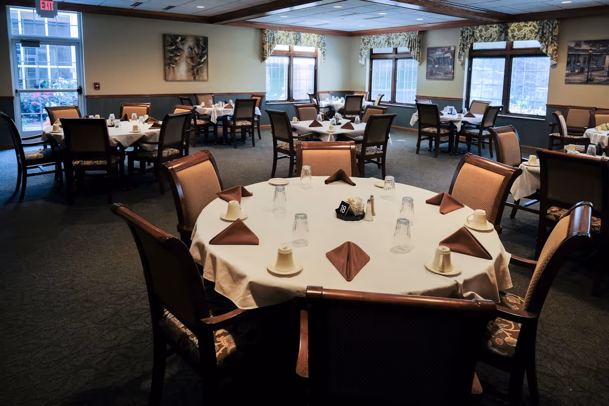 Dining room with multiple round tables set with white tablecloths, folded napkins, cups and chairs beneath large windows.