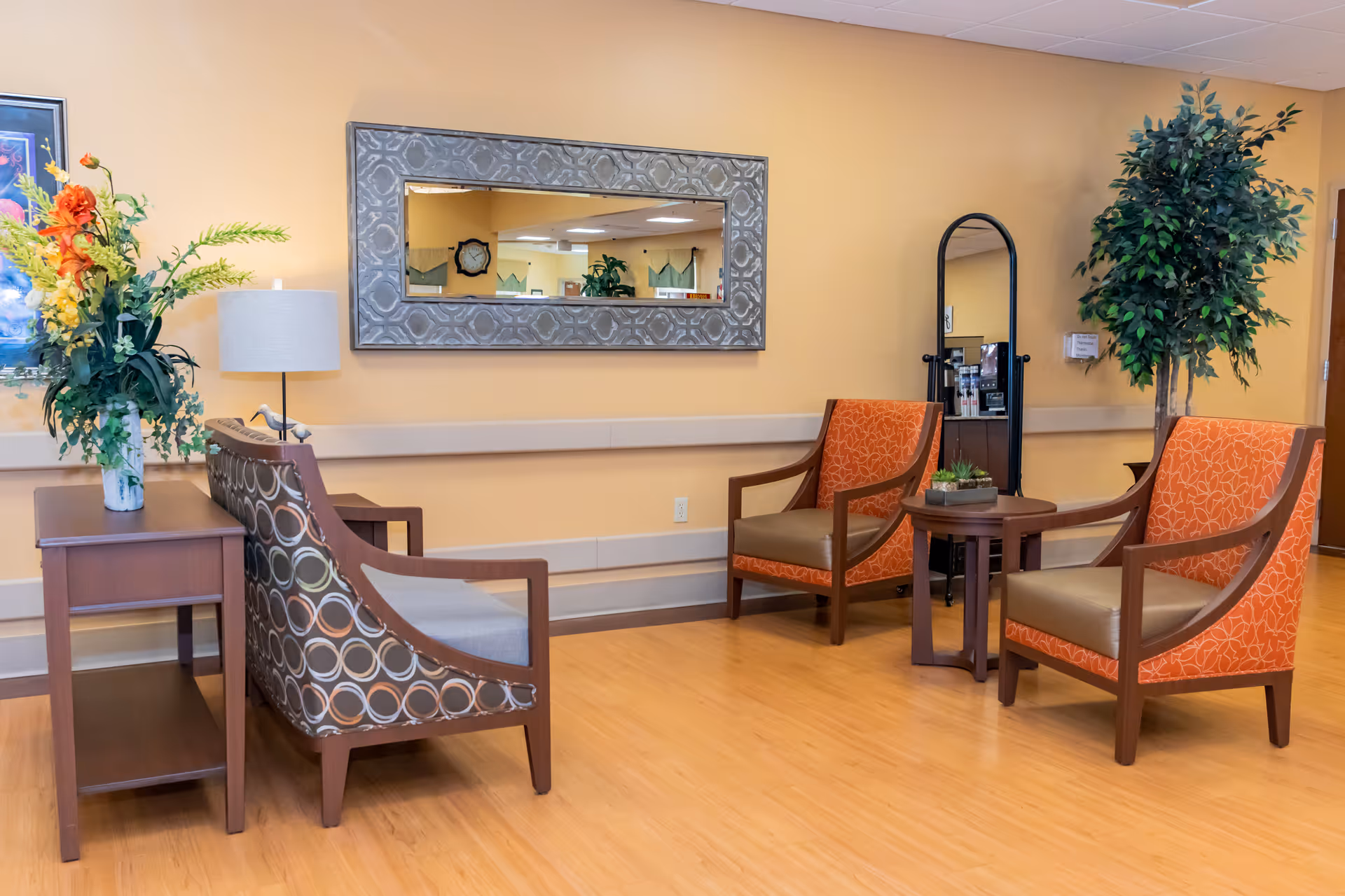 A cozy seating area in a senior living facility with two orange patterned armchairs, a small round wooden table with a plant, a patterned loveseat, a wooden side table with a lamp and a vase of flowers, a large decorative mirror on the wall, and a tall artificial plant in the corner.