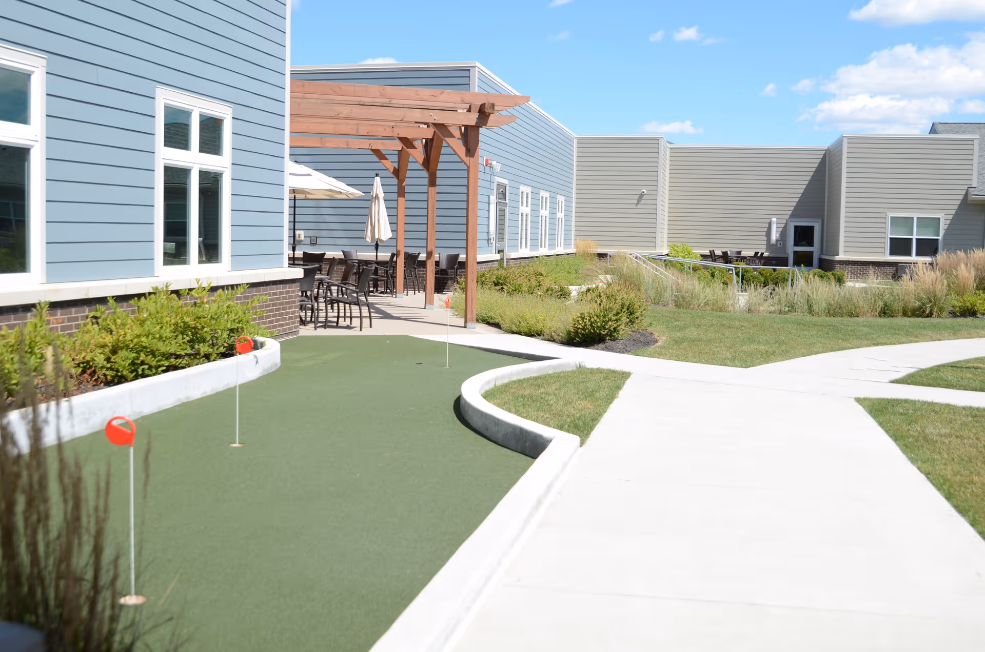 Outdoor area of a senior living facility featuring a small putting green with red flags, a concrete walkway, landscaped greenery, and a patio with tables, chairs, and umbrellas under a wooden pergola. The building exterior is clad in light blue and beige siding with white-framed windows under a clear blue sky.