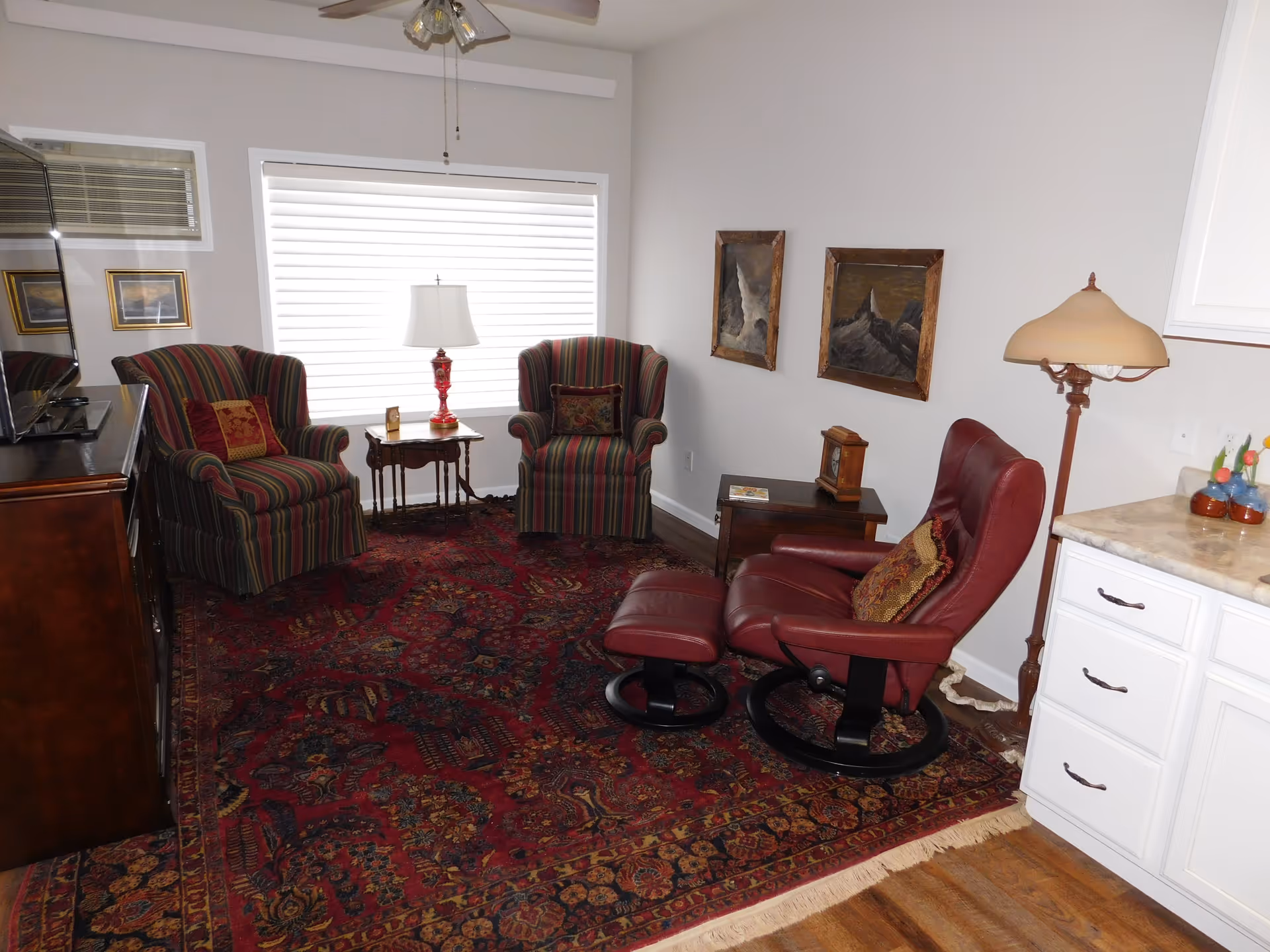 A cozy living room with a large patterned red rug covering the wooden floor. There are two striped armchairs with decorative pillows facing a TV on a wooden cabinet. A small wooden side table with a lamp is placed between the armchairs. A red leather recliner with a matching footrest and a decorative pillow is positioned near a floor lamp and white cabinetry. Two framed paintings hang on the wall.