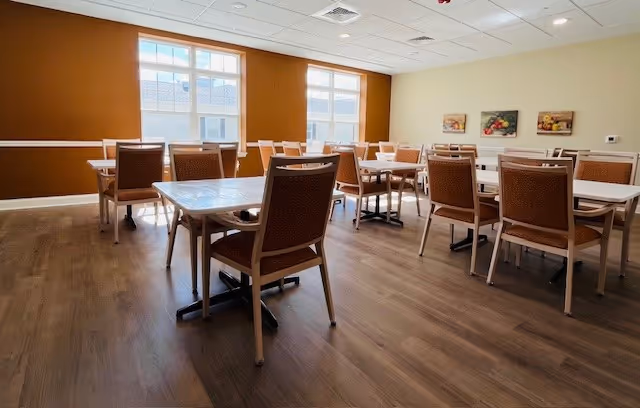 Bright communal dining room with multiple tables and chairs, large windows, warm orange accent wall, and wood-look flooring.