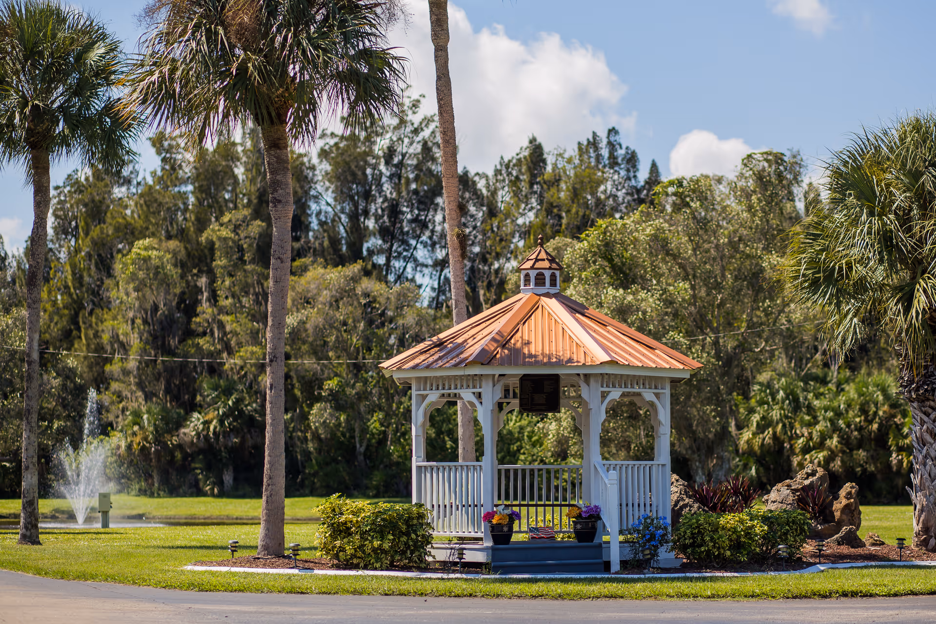 A white gazebo with a copper-colored roof surrounded by green grass, palm trees, and bushes under a partly cloudy blue sky. A water fountain is visible in the background.