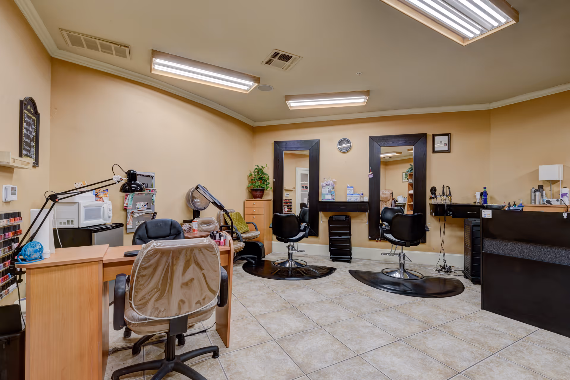 Interior view of a salon area in a senior living facility with two black salon chairs in front of large mirrors, a manicure table with a chair covered in plastic, nail polish racks, a microwave on a small refrigerator, and various salon equipment and supplies.