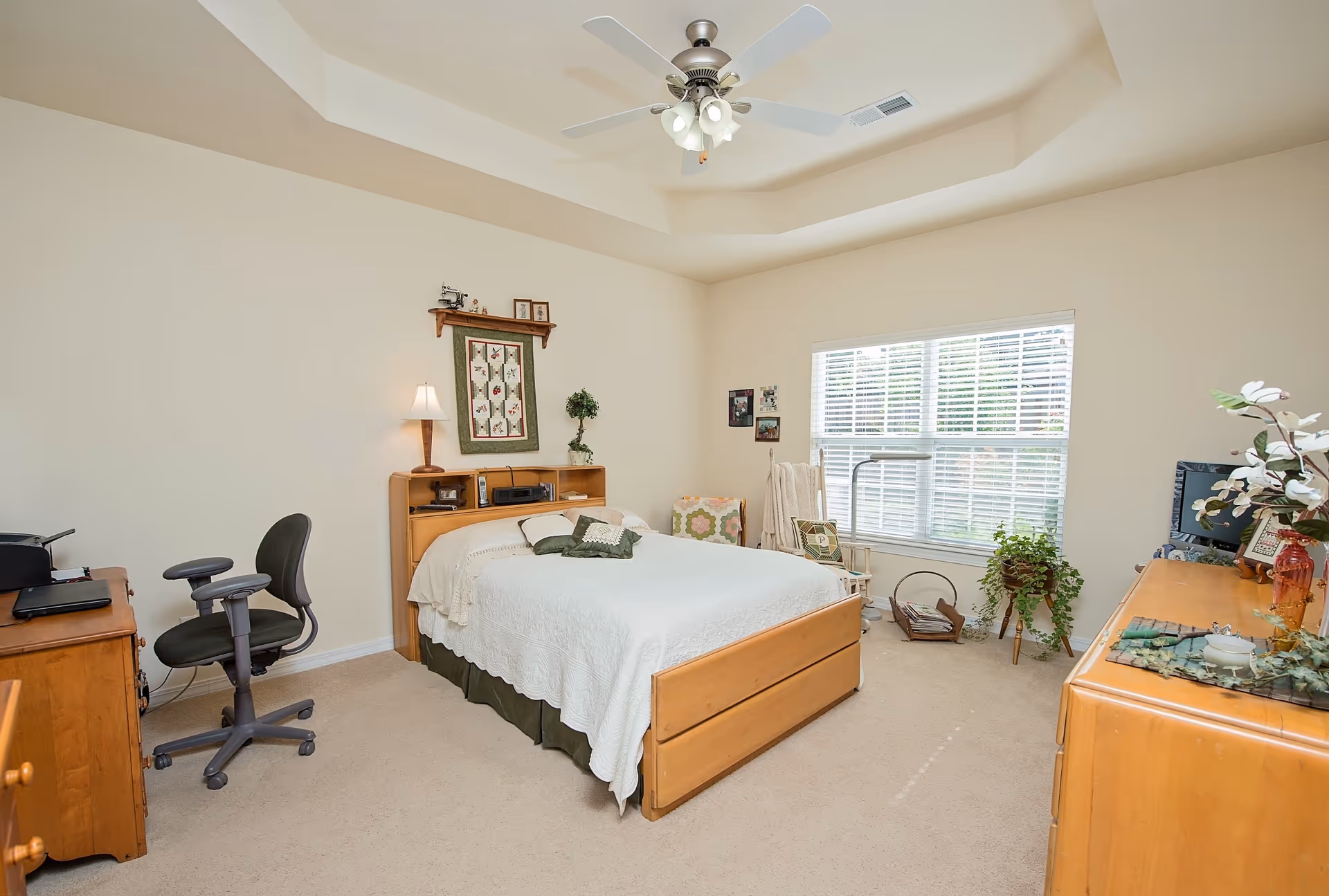 A bright and cozy bedroom with a wooden bed covered in white bedding and green pillows. The room features a ceiling fan with lights, a wooden dresser with decorative items, a desk with an office chair, a window with white blinds, and various plants and framed pictures on the walls.