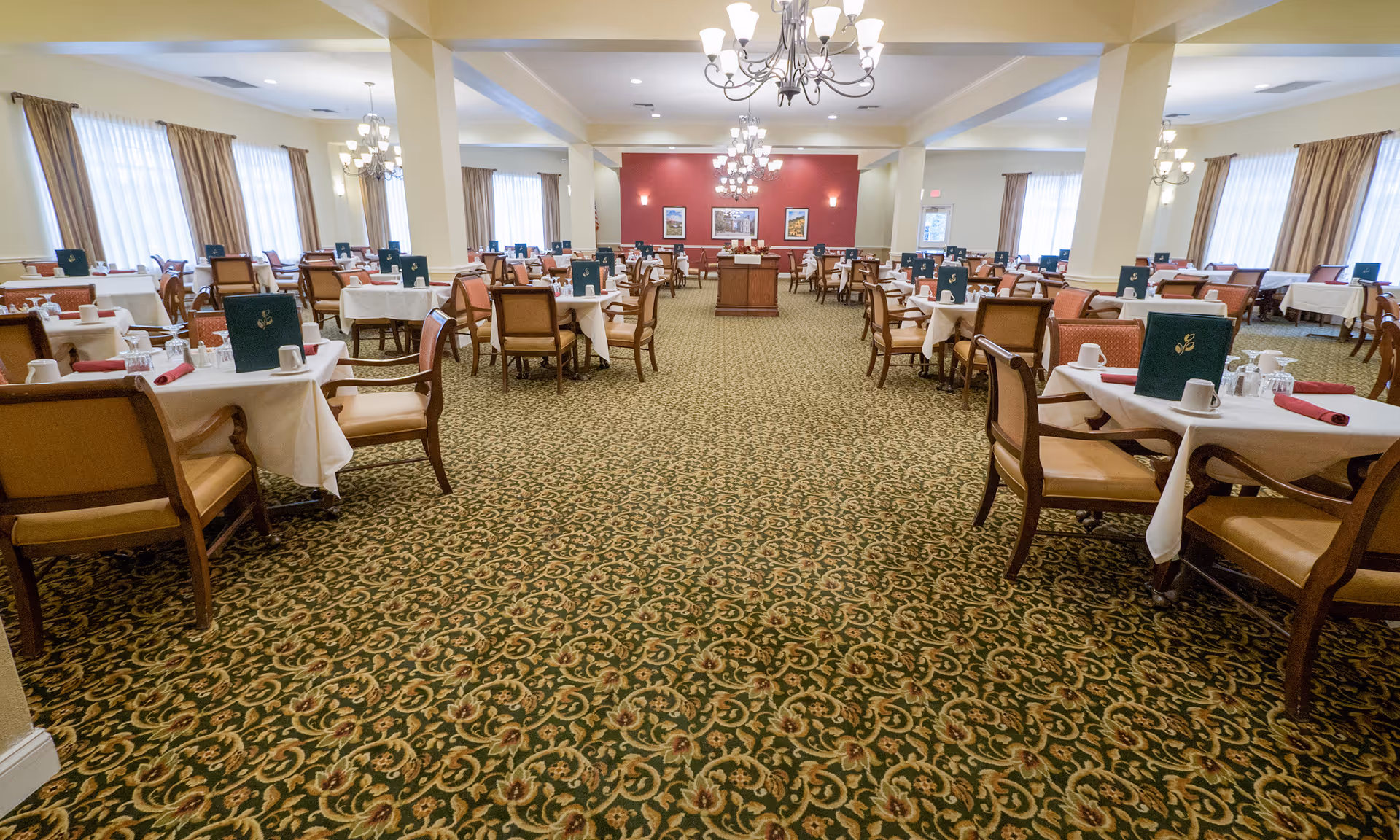 A spacious dining room with multiple tables covered in white tablecloths, each set with cups, glasses, and folded red napkins. The room has patterned carpet flooring, large windows with curtains, and chandeliers hanging from the ceiling. The walls are painted in light colors with one accent wall in red at the far end.