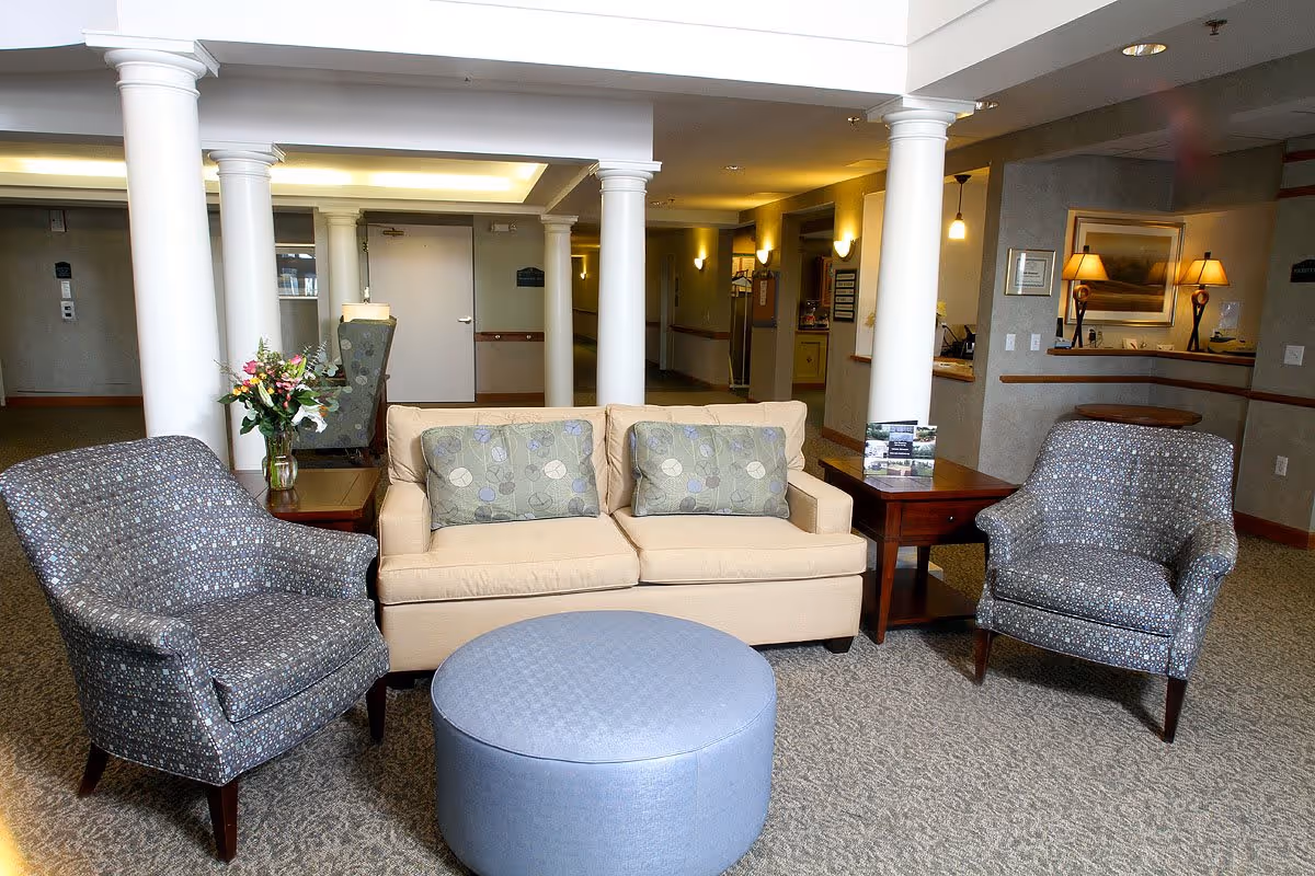 A cozy seating area in a senior living facility featuring a beige sofa with two patterned cushions, two patterned armchairs, a round blue ottoman, and two wooden side tables. One side table has a vase with flowers, and the other has brochures. The background shows white columns, a hallway, and a reception desk with warm lighting.
