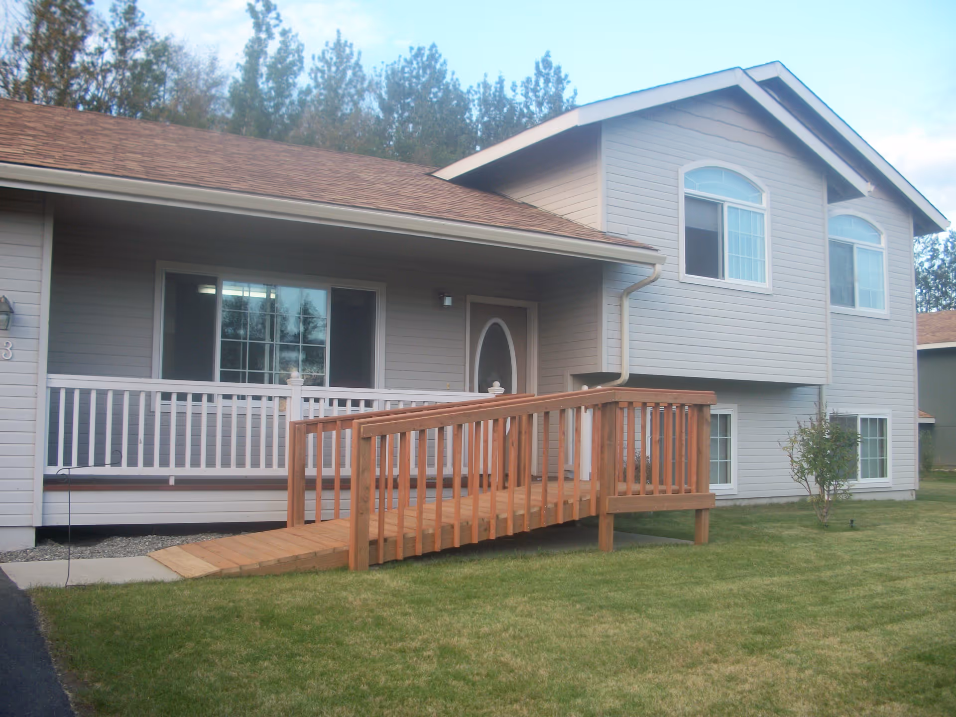 Exterior view of a two-story residential building with light gray siding and a brown shingled roof. There is a wooden wheelchair ramp leading up to a covered porch with white railing. The lawn is green and well-maintained, and there are trees in the background.