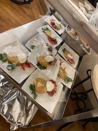 Several plates of dessert arranged on a metal cart, each plate containing a pastry topped with whipped cream and garnished with strawberries and a mint leaf, covered partially with plastic wrap.