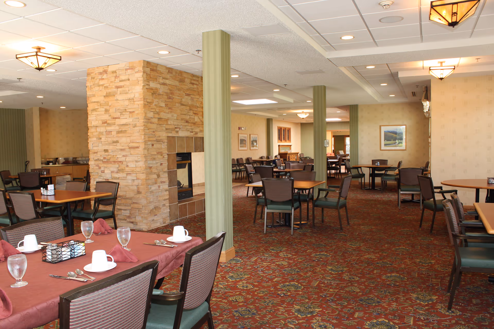 A spacious dining room with multiple tables and chairs arranged neatly. The tables are set with glasses, cups, napkins, and cutlery. The room features a carpet with a floral pattern, a stone fireplace in the center, and soft lighting from ceiling fixtures. The walls are decorated with framed pictures and light-colored wallpaper.