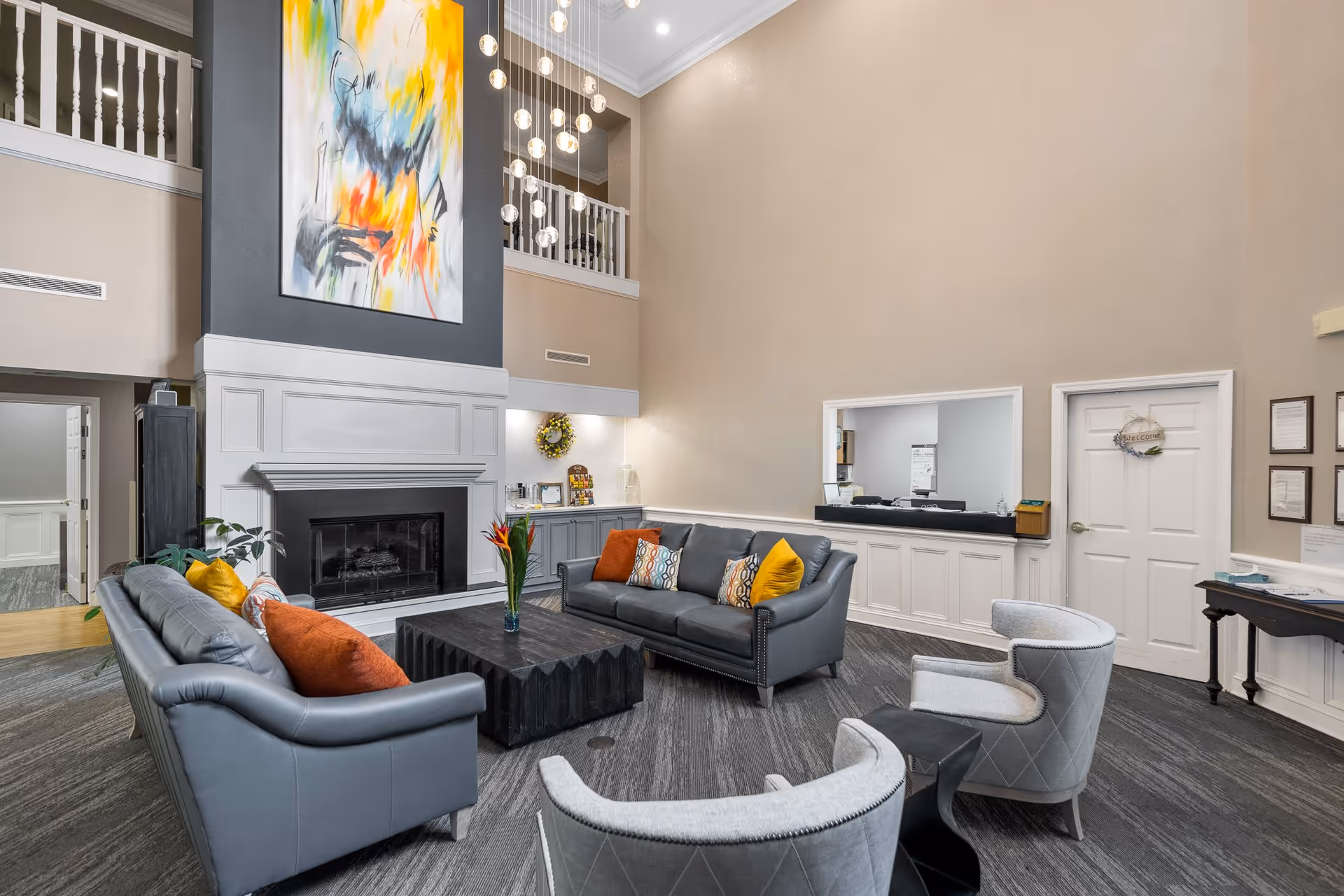 A spacious and well-lit living room area with high ceilings featuring two gray leather sofas with colorful throw pillows, two gray upholstered armchairs, and a black coffee table with a vase of flowers. There is a large modern abstract painting above a white fireplace, and a contemporary chandelier with multiple hanging glass orbs. The room has beige walls, gray carpet flooring, and a reception window with a door labeled 'Welcome' nearby.