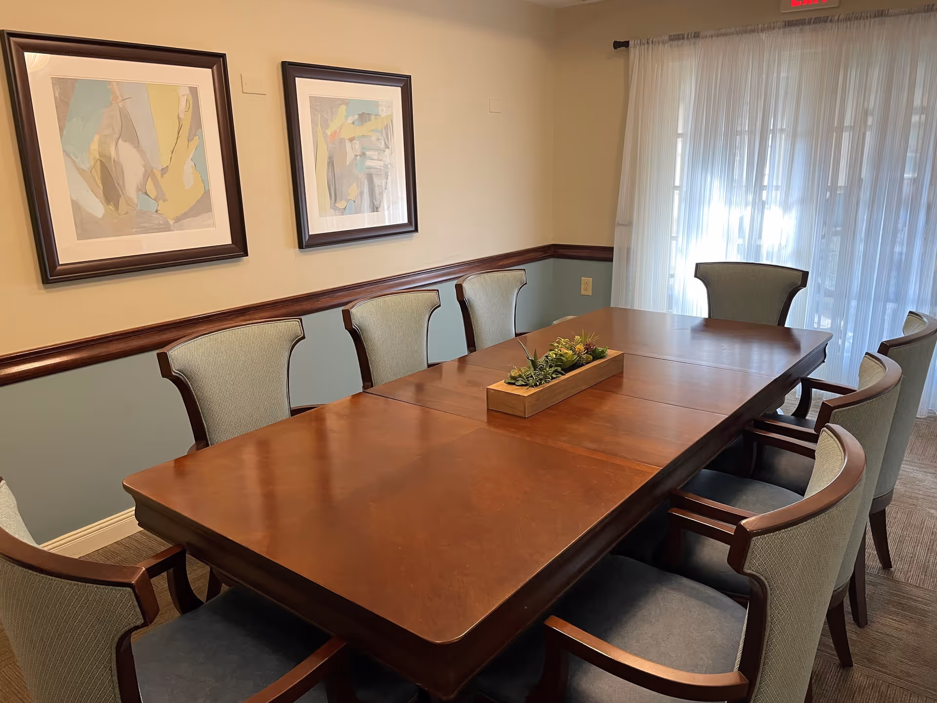 A dining room with a large wooden table surrounded by eight upholstered chairs. The table has a rectangular wooden planter with green plants in the center. Two abstract paintings hang on the wall, and sheer white curtains cover a large window or door letting in natural light.