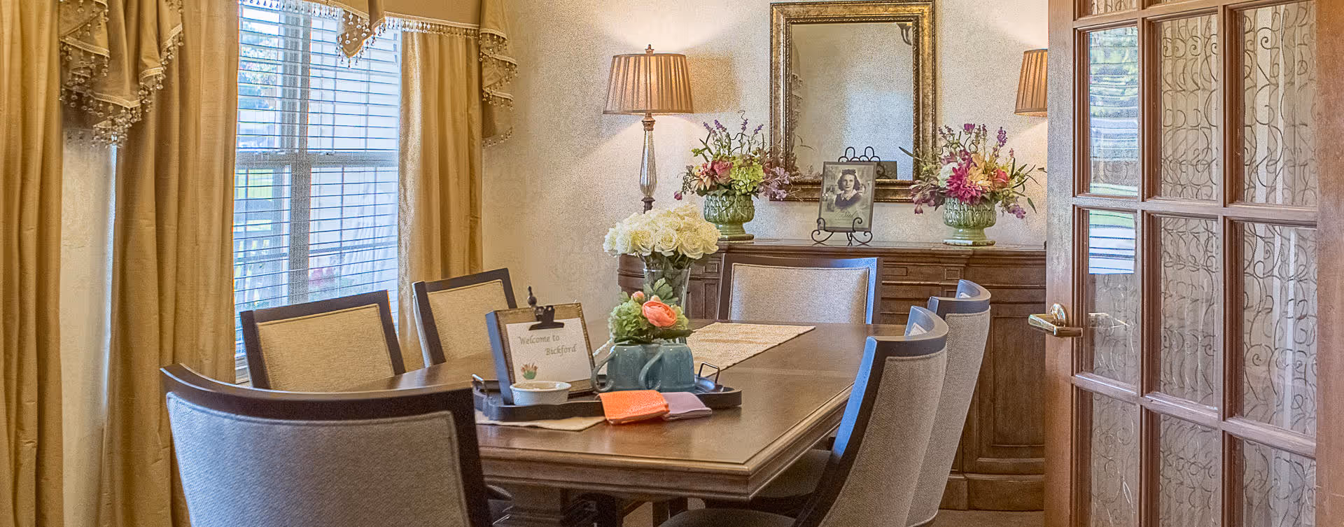 A warmly decorated dining room with a wooden dining table surrounded by eight upholstered chairs. The table has a centerpiece with flowers and a small tray with a welcome sign. There are large windows with beige curtains on the left, a wooden sideboard with floral arrangements and a framed photo, and a mirror on the wall. A lamp with a beige shade is also visible on the sideboard. The room is viewed through a glass-paneled wooden door on the right.