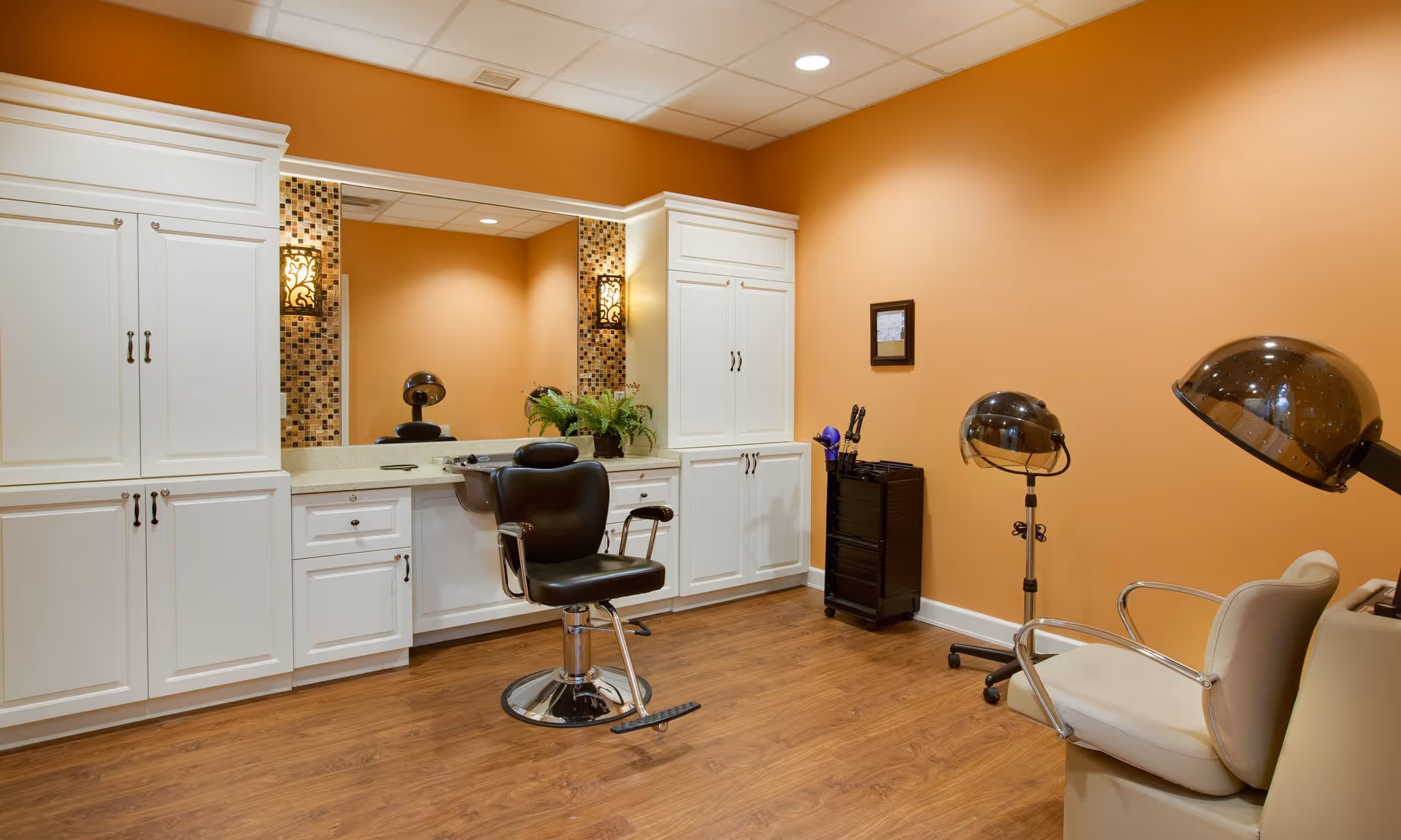Interior view of a salon area in a senior living facility with a black salon chair in front of a large mirror, white cabinetry, wooden floor, and two hair drying stations with dome dryers.