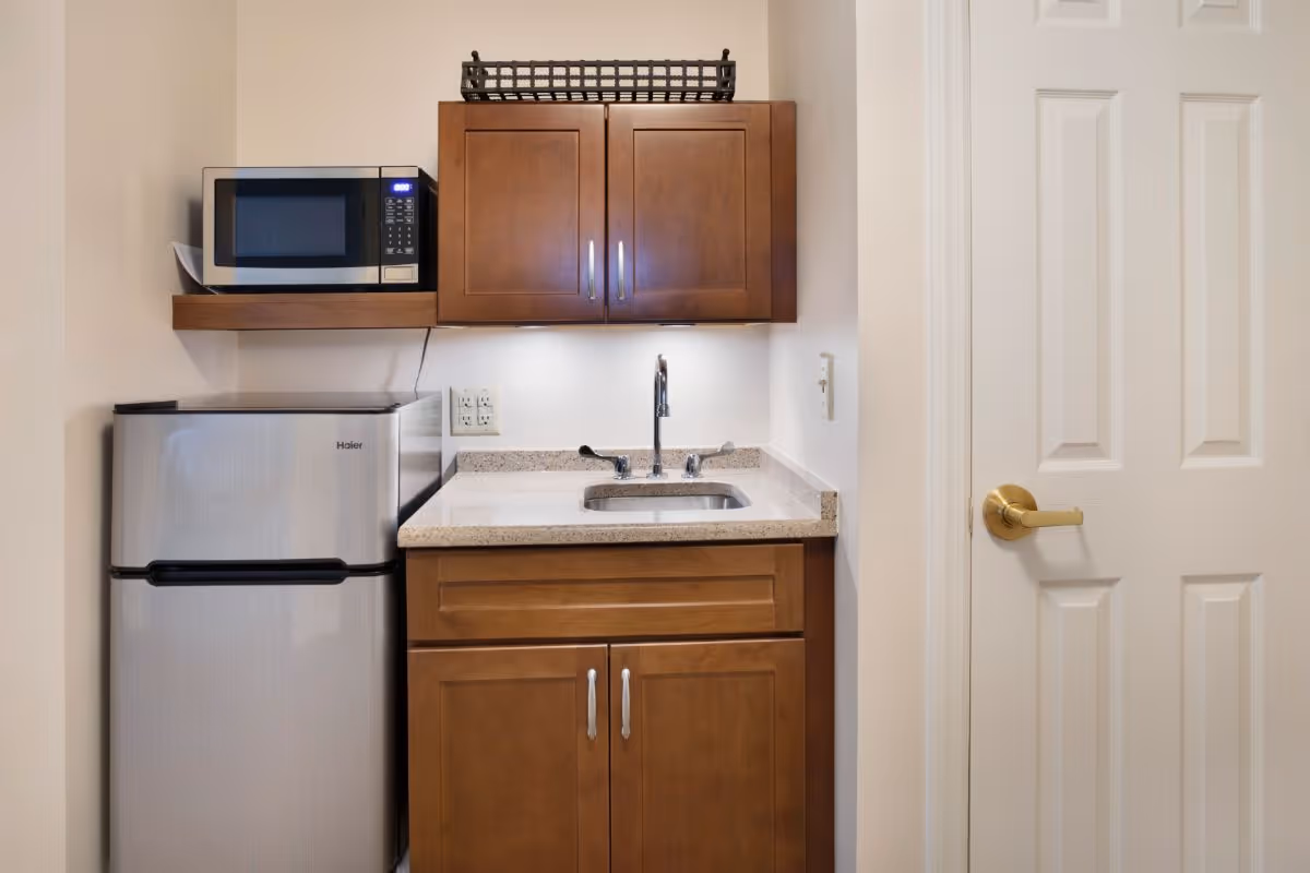 A small kitchenette area featuring a stainless steel mini refrigerator, a microwave on a wooden shelf above it, wooden cabinets above and below a countertop with a built-in sink and faucet, and a white door with a gold handle to the right.