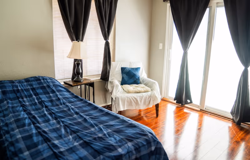 A tidy bedroom with a bed, plaid blanket, white chair, lamp, and sliding glass doors.