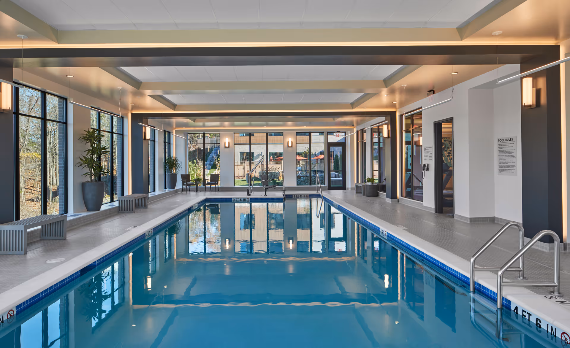 Modern indoor swimming pool with large windows, benches, potted plants, and overhead lighting.