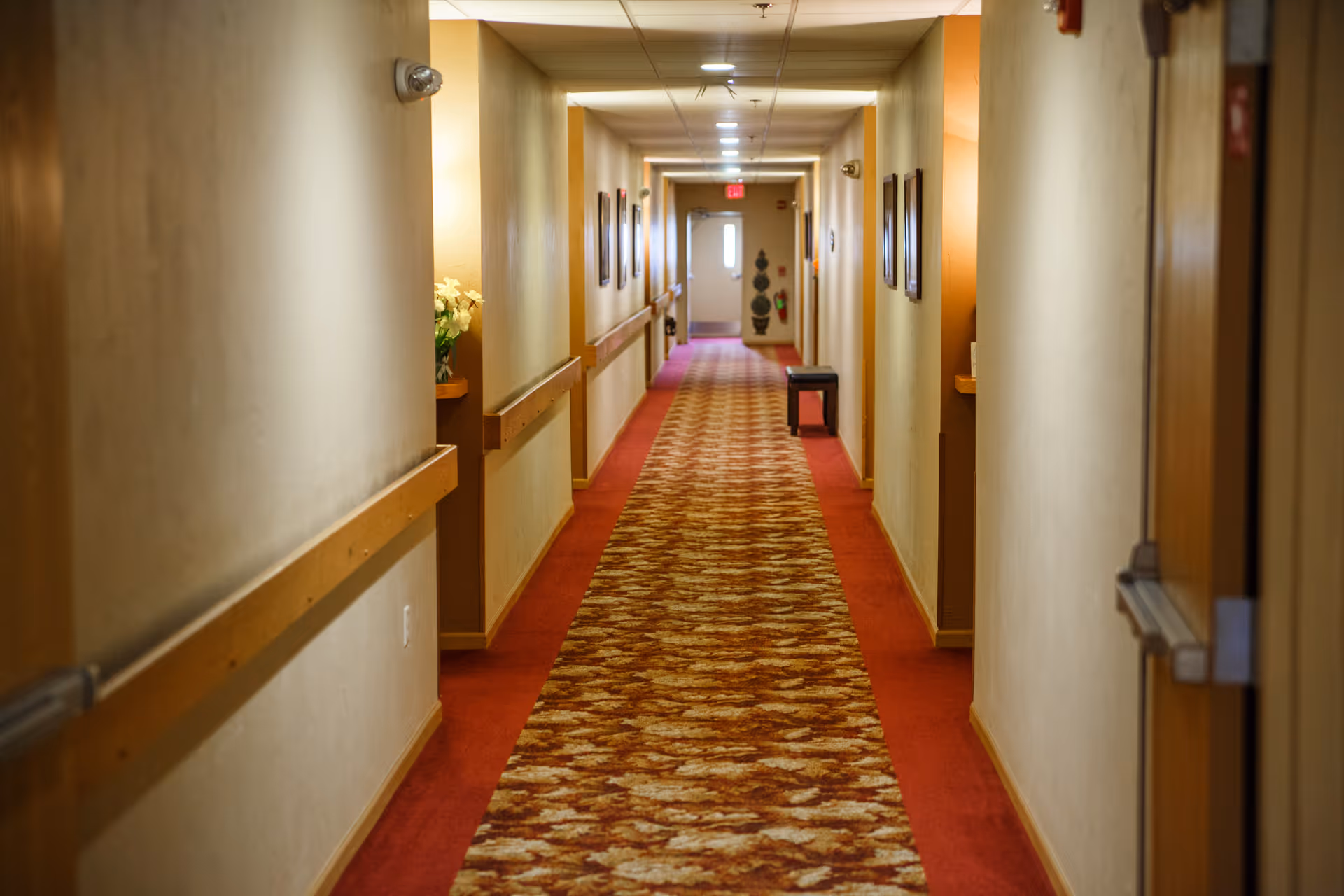A long, narrow hallway in a senior living facility with beige walls, wooden handrails on both sides, and a patterned carpet runner in shades of red and brown. The hallway is lit with ceiling lights and has framed pictures on the walls. A small bench is positioned along the right side near the end of the hallway, and a door with a window is visible at the far end.