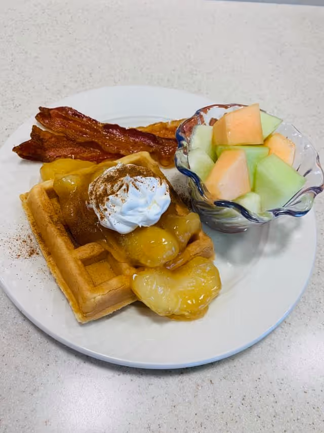 A plate with a waffle topped with fruit compote and whipped cream, two strips of bacon, and a small glass bowl of melon cubes on a countertop.