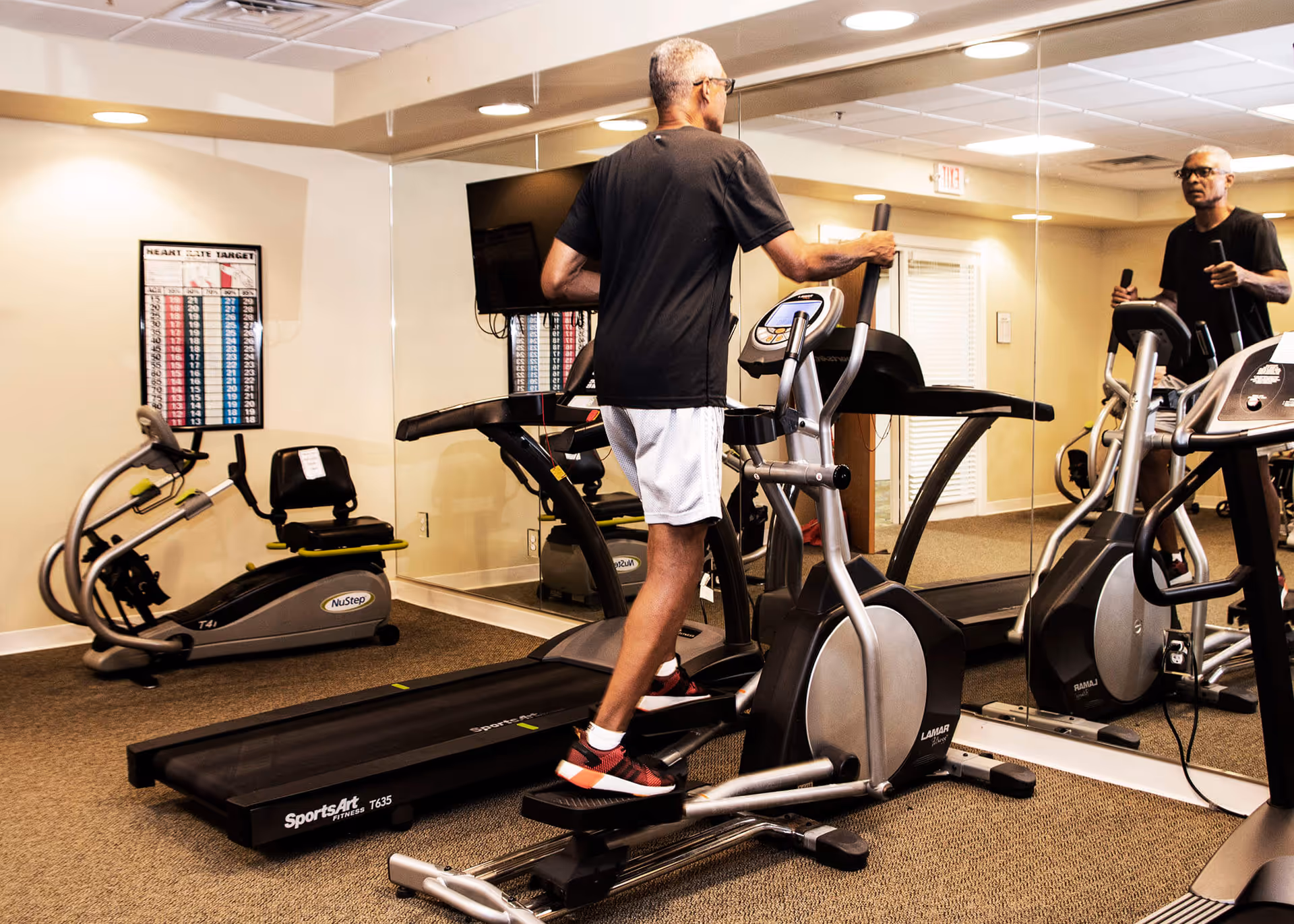 A man exercising on an elliptical in a mirrored fitness room with treadmills and a recumbent bike.